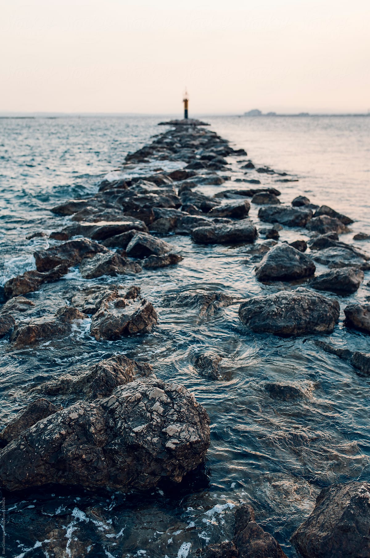 A lighthouse at sunset surrounded by water and rocky path, out of focus.