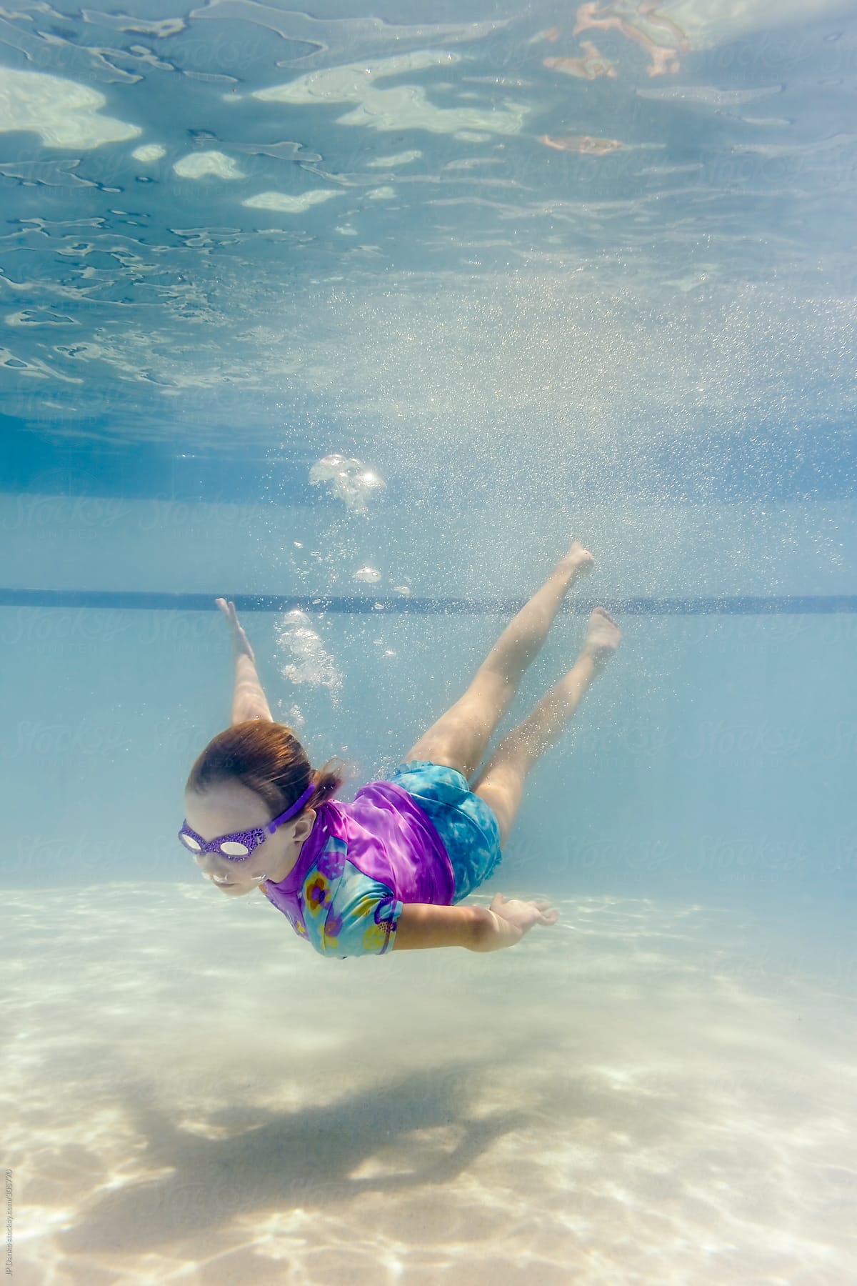Little Girl Swimming Underwater In Swimming Pool Wearing Goggles By