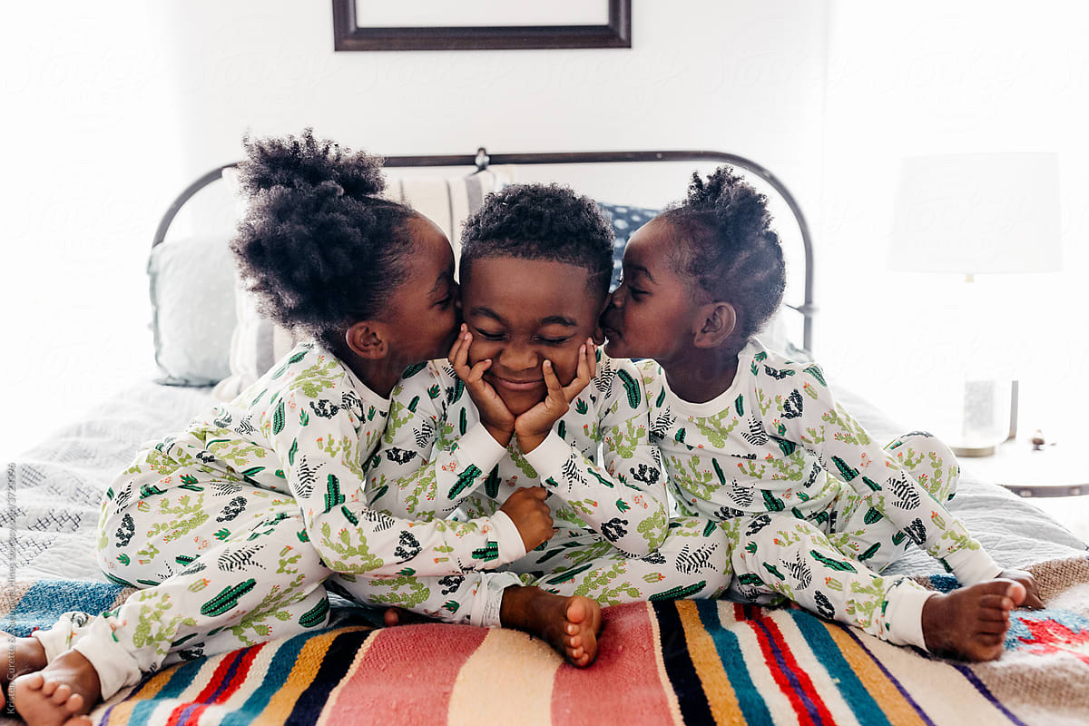 Three siblings in matching pajamas sitting on the bed. The two sisters are kissing their brother.
