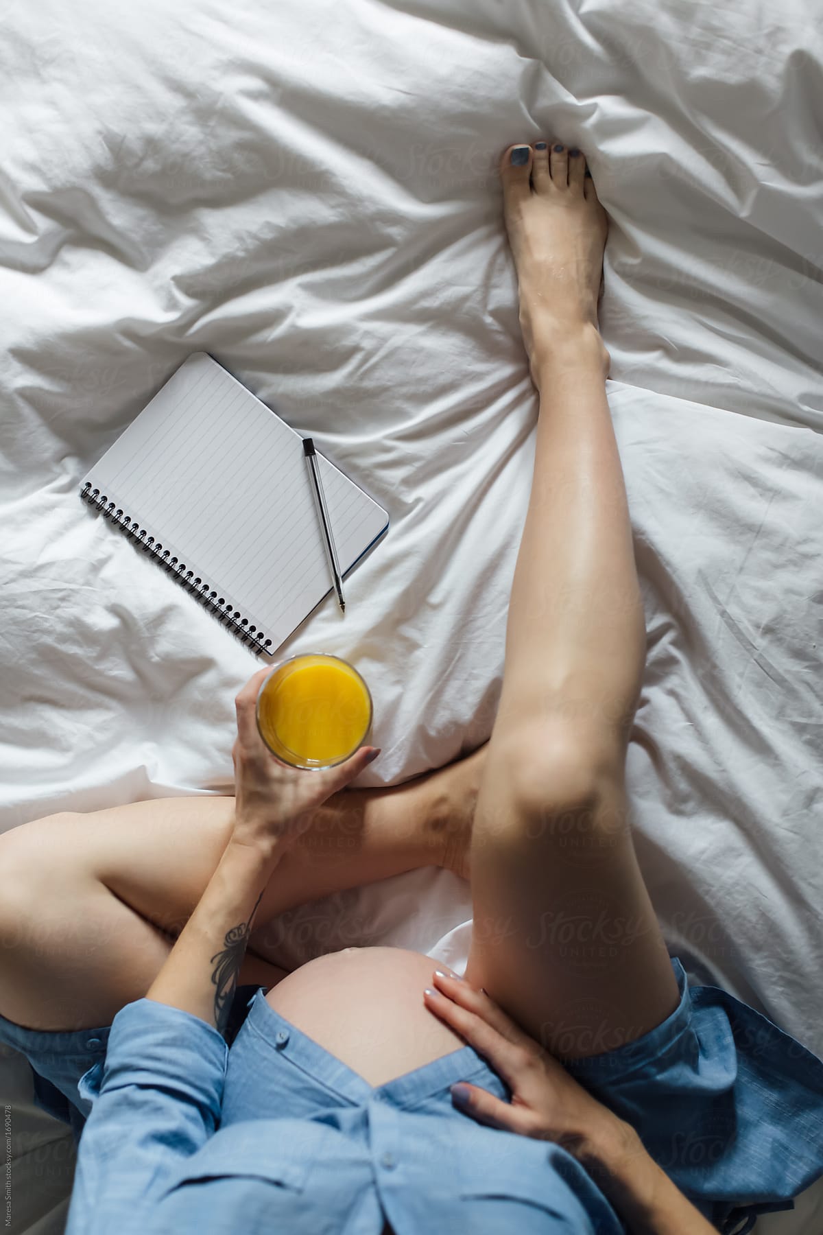 A pregnant women in a blue shirt with bare legs and bump, holding a glass of orange juice with a blank notebook on the bed