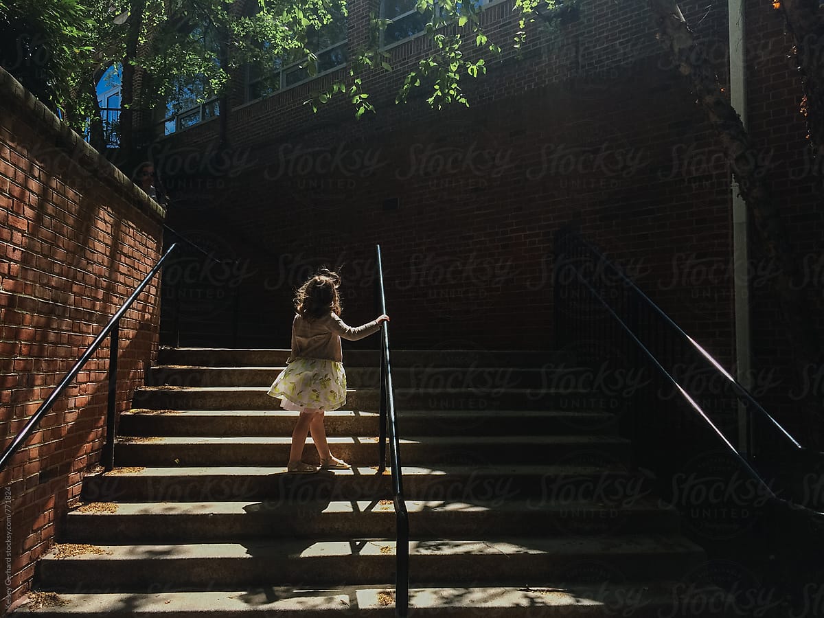 A young girl in a dress stands in a shadowy, light-filled stairwell outdoors.