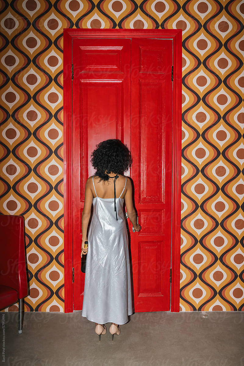 A curly-haired woman in a silver dress stands in front of a red door