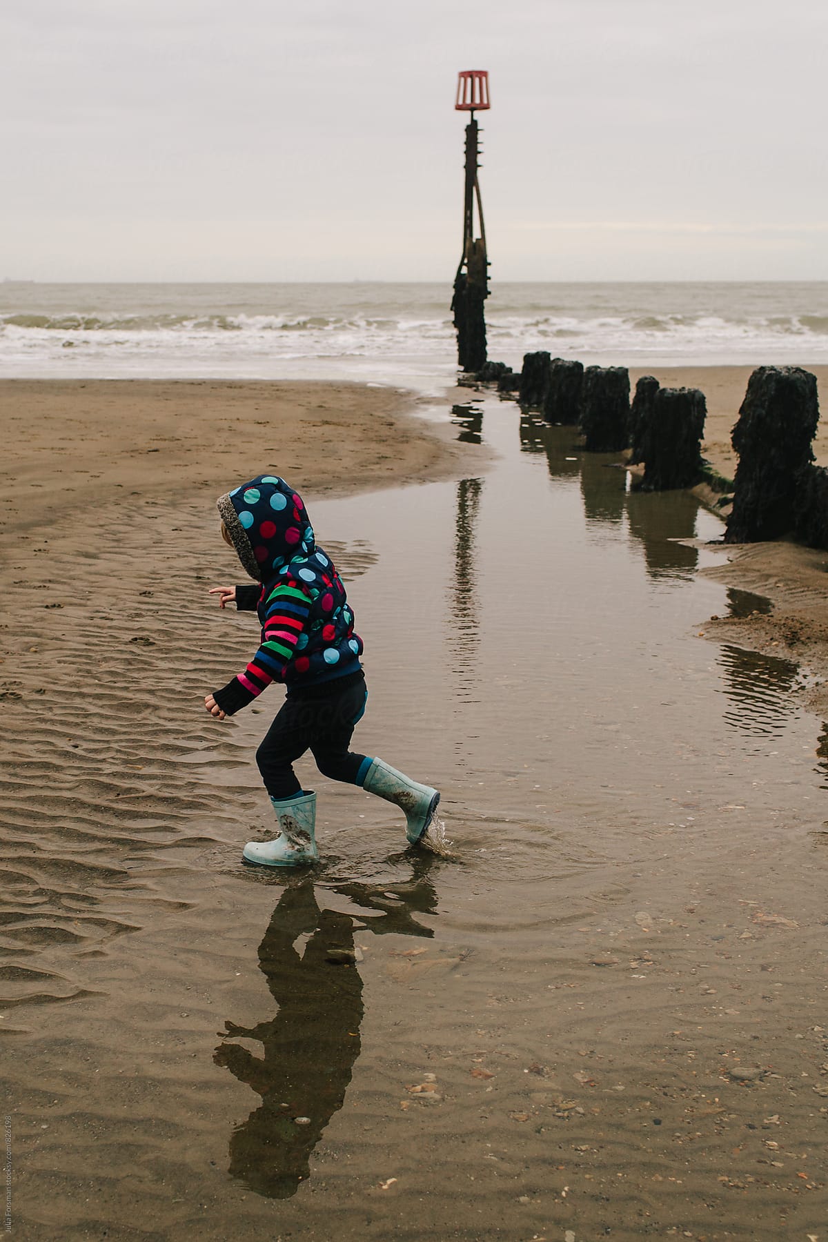 Small child and her reflection running through water next to a sea barrier on a wintry beach.
