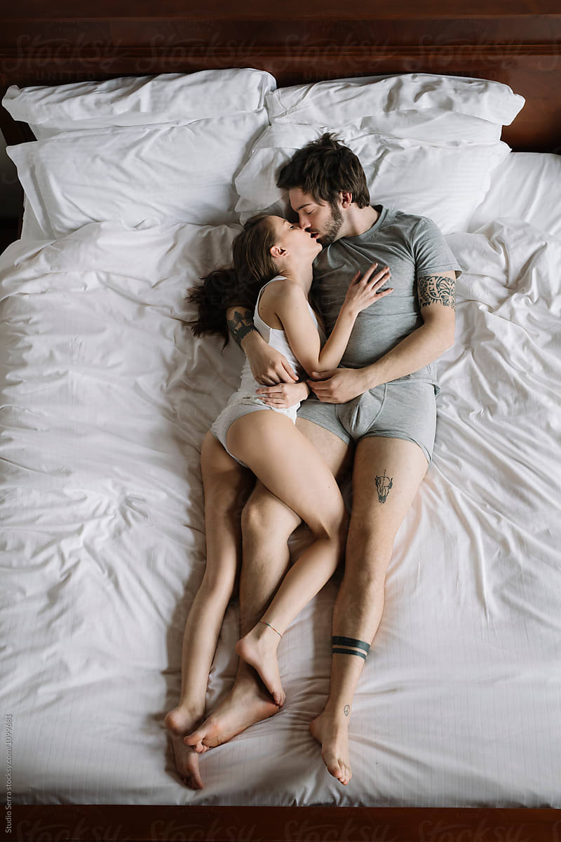 Overhead of young couple kissing on duvet on bed in morning light.