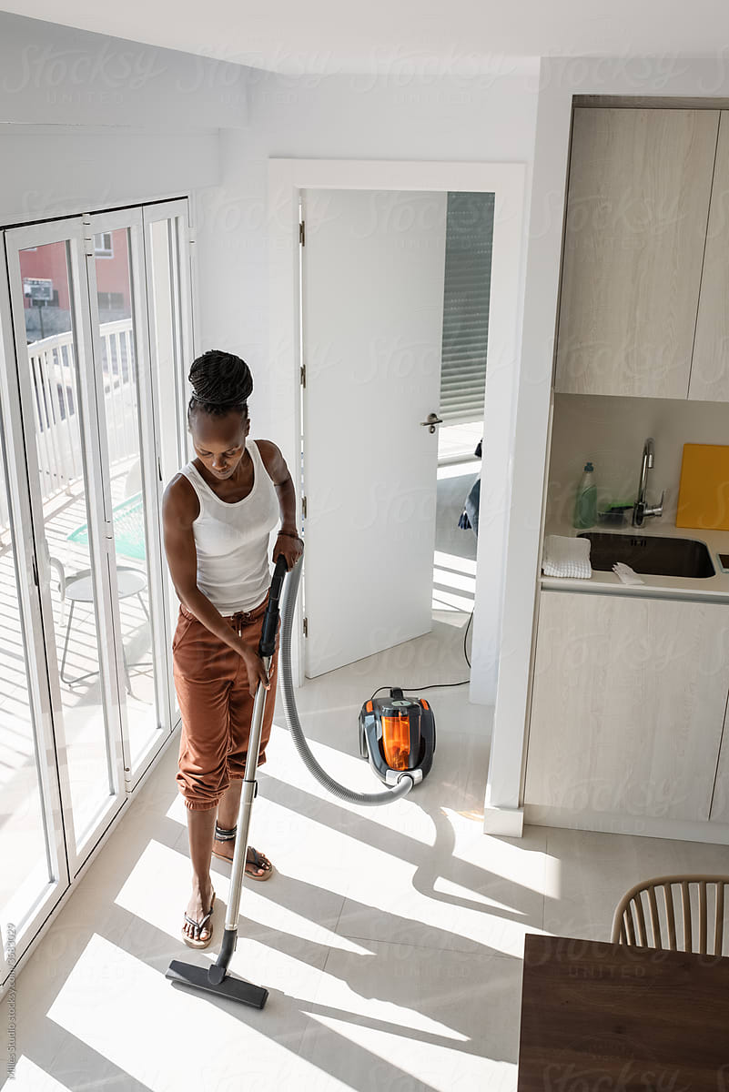 Black housewife vacuuming floor near balcony door