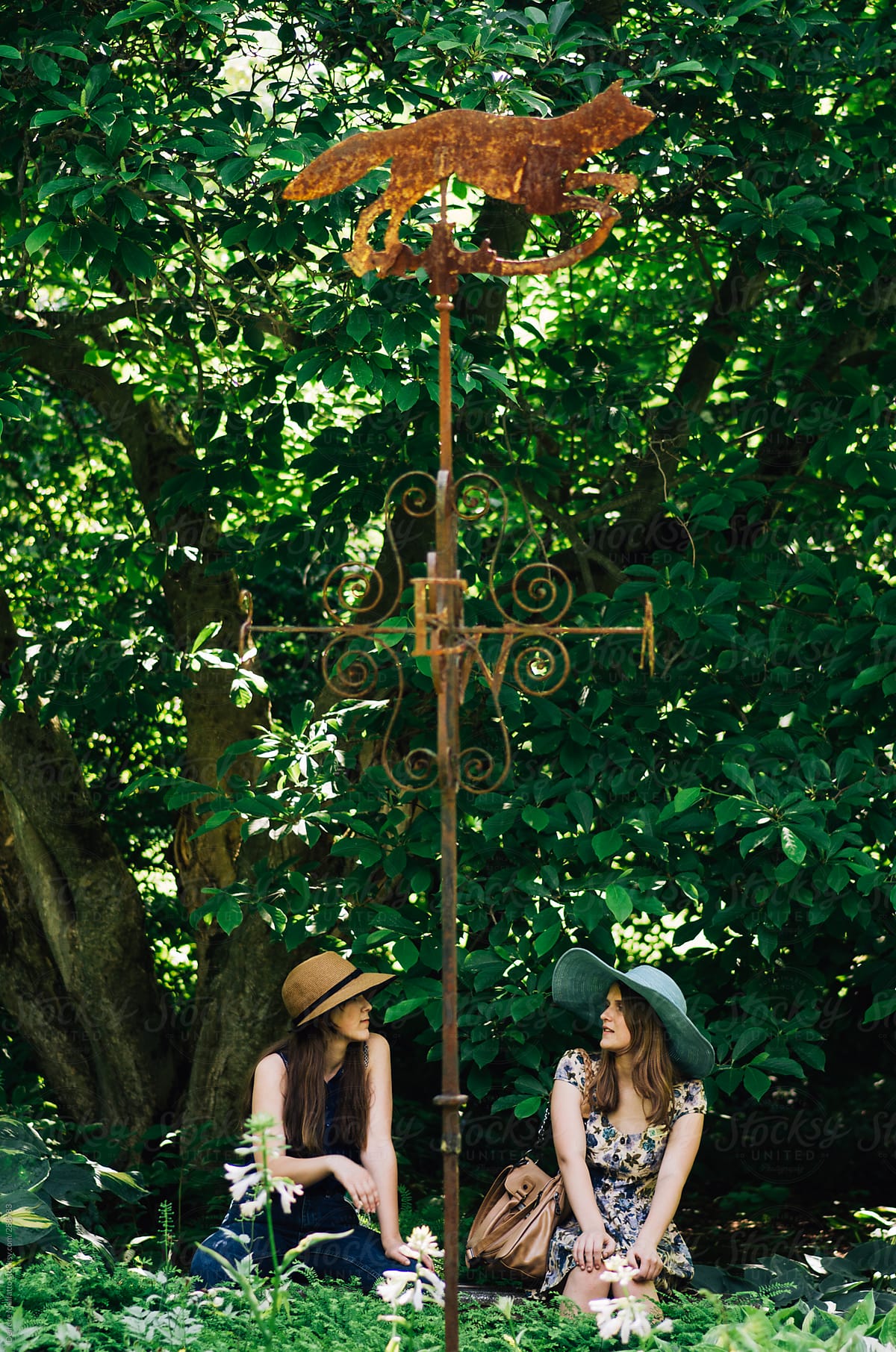 two young woman in hats talk in a garden under a weathervane