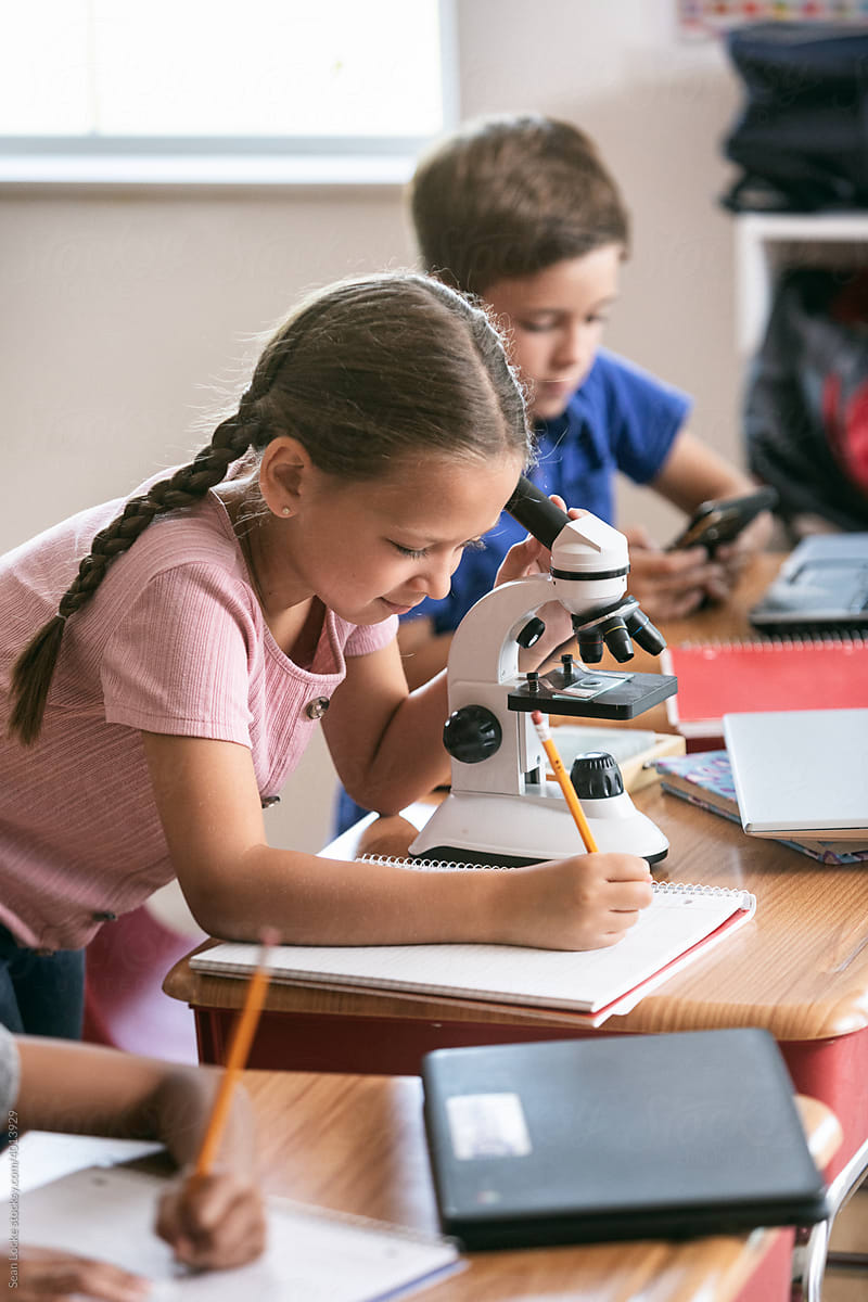 School: Female Student Using Microscope And Taking Notes