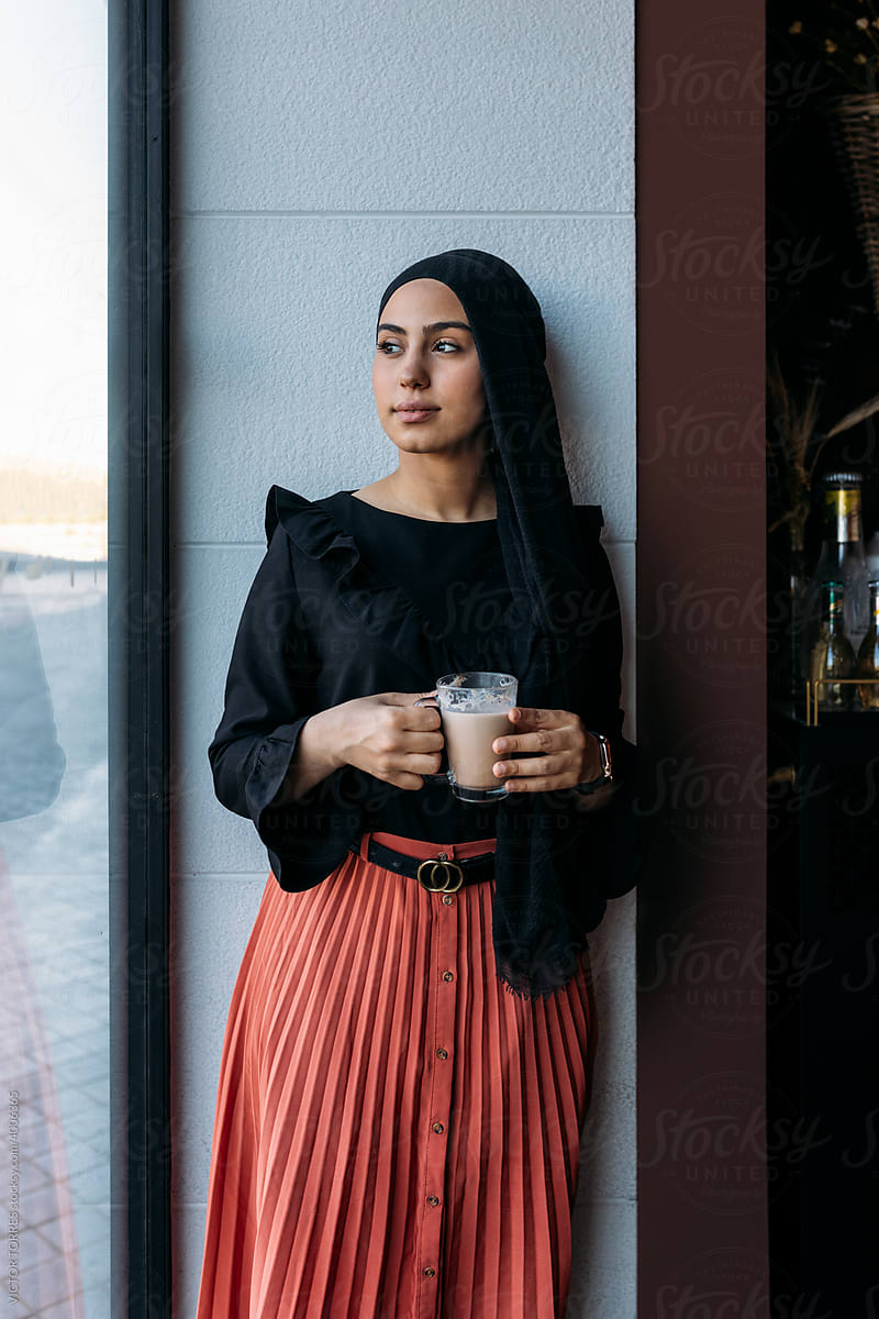 Pensive muslim woman in headscarf standing besides a window