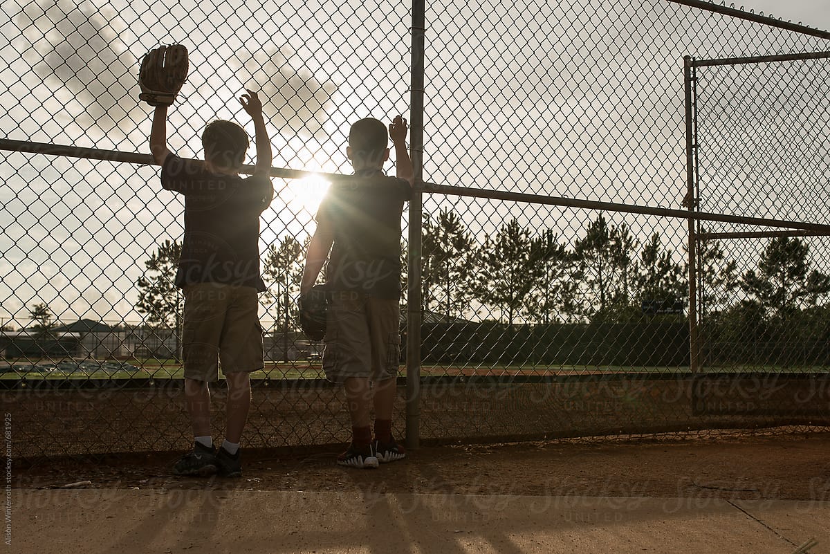 Two Boys Stand On A Ball Field At The Chain Link Fence