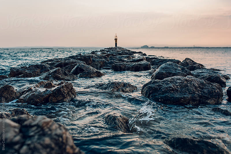 A lighthouse at sunset surrounded by water and rocky path, out of focus.