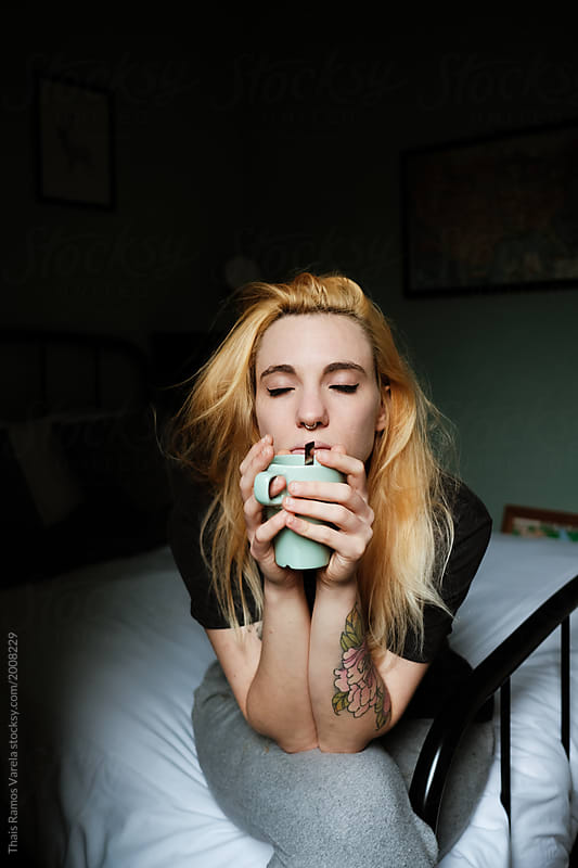 sleepy blonde woman sitting on the bed taking a cup of coffee in the morning