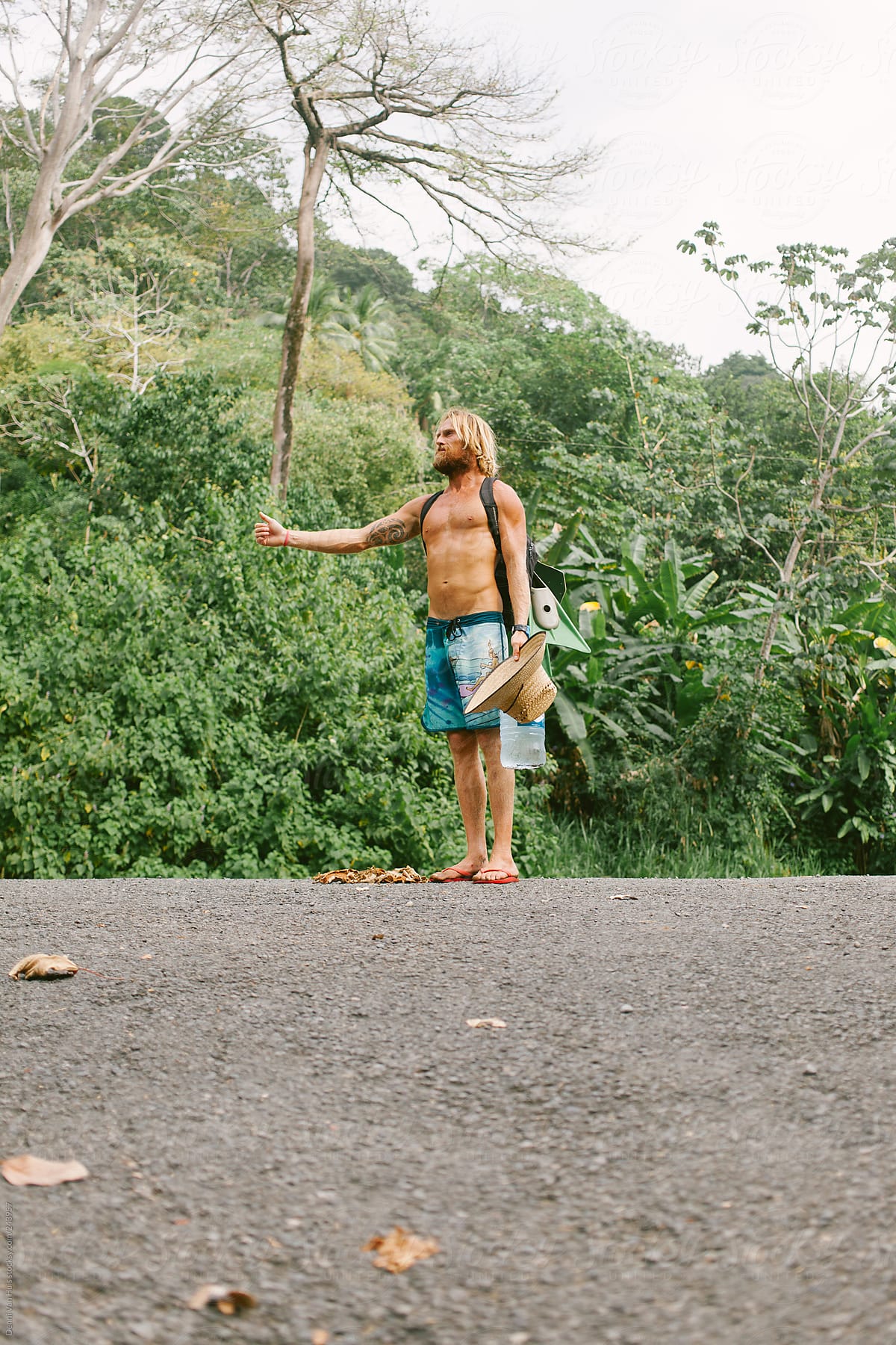 Man with no shirt on hitchhiking on the side of the road.
