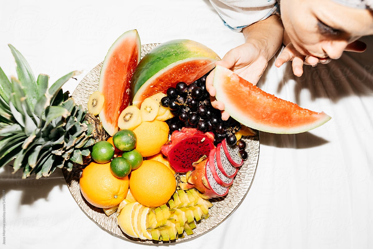 large fruit tray on bed with woman eating watermelon shot from above