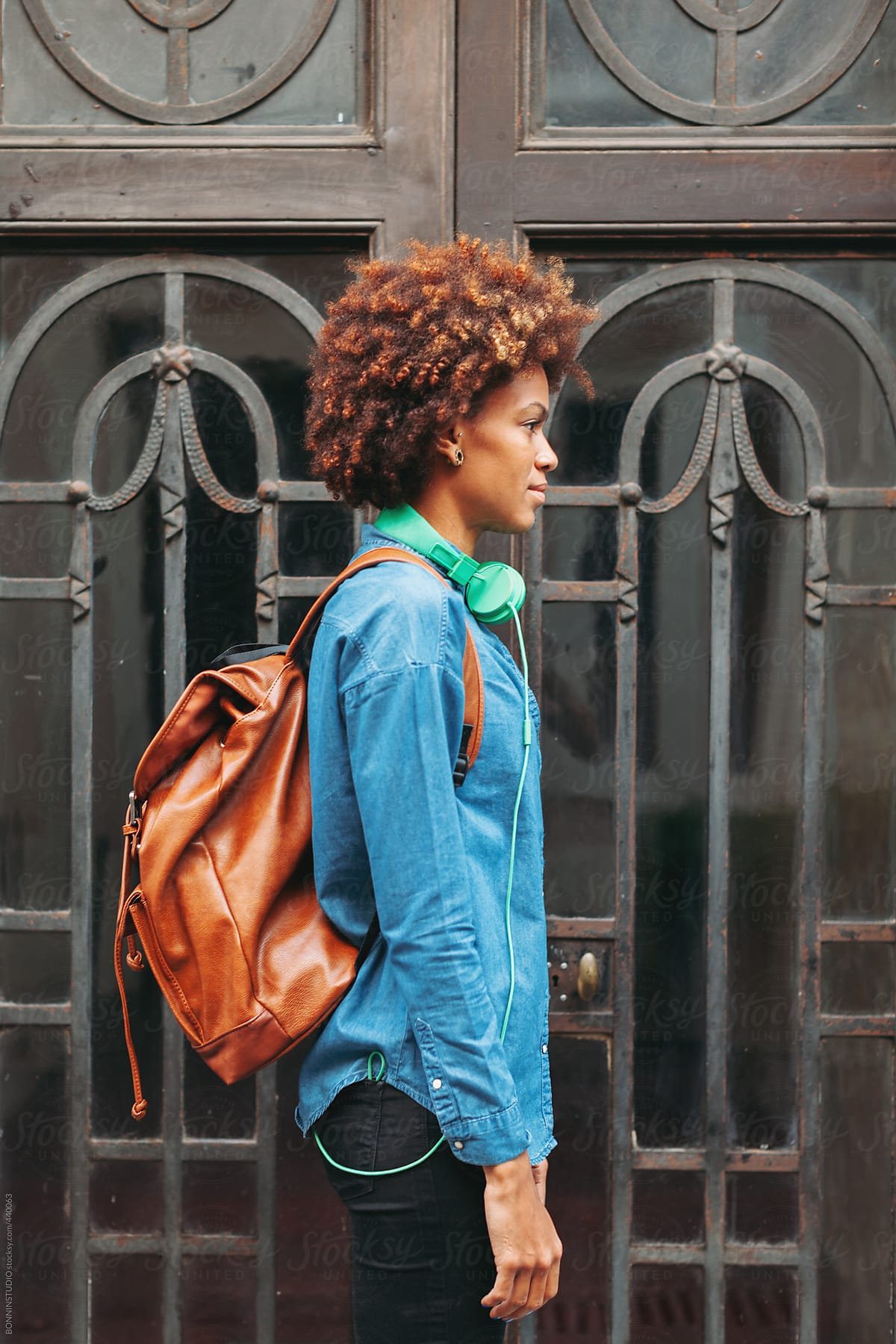 Profile portrait of afro american woman with leather backpack and green headphones in outdoors.
