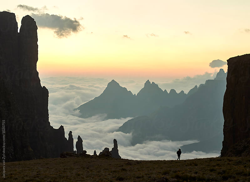 A hiker silhouetted and spire rock faces in an epic landscape with sharp mountain tops breaking through the clouds at sun rise.