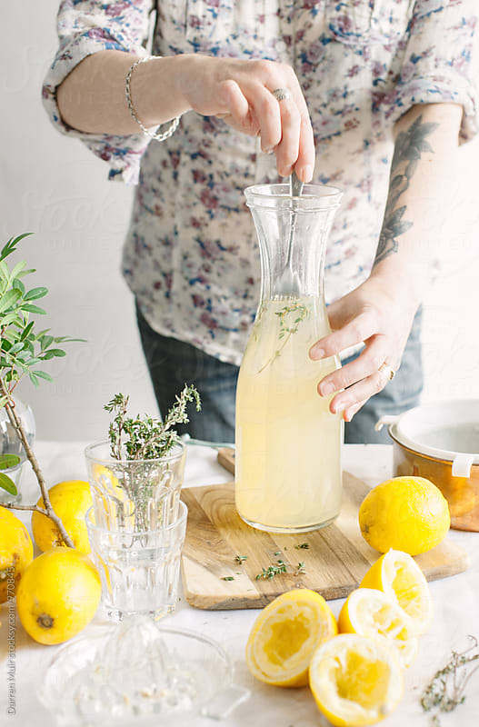 Series showing the making of homemade lemonade infused with thyme: Woman stirring thyme sprigs through a carafe of homemade lemonade.