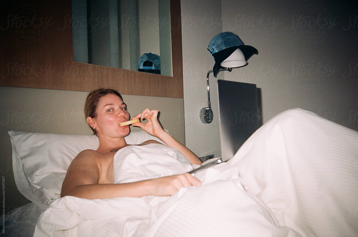 Naked woman in white blanket lying on bed while eating