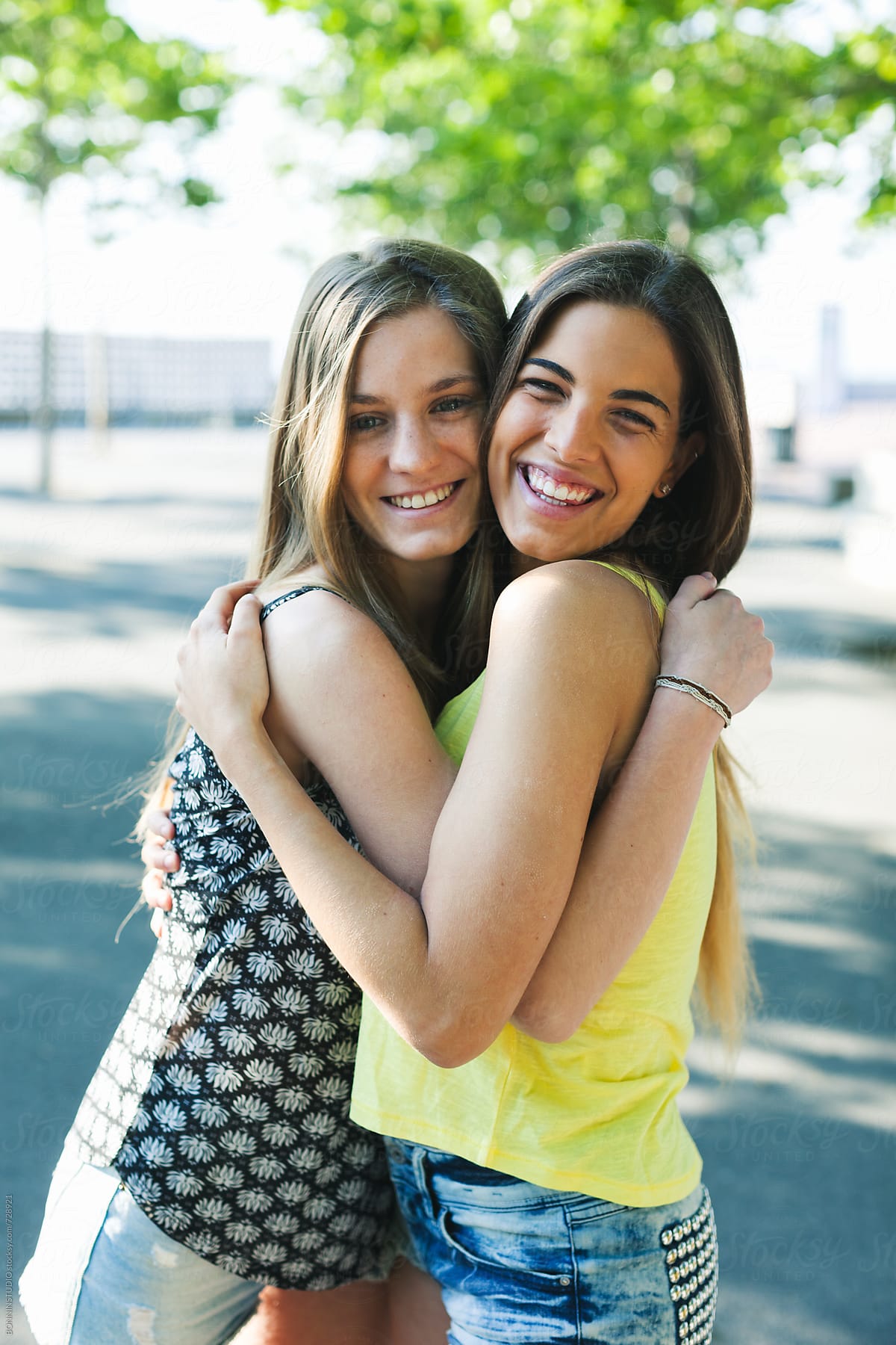 "Young Female Friends Embracing Together Standing In A Park." by