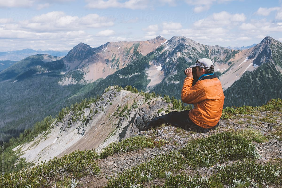 Man Sitting On Ledge, Looking Through Binoculars At Vast Mountain Range by Rialto Images