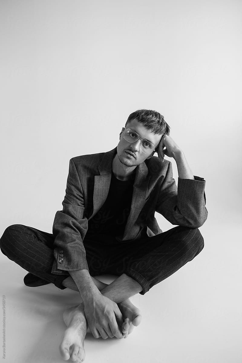 black and white portrait of a guy in stylish glasses and a jacket sitting on the floor in a relaxed pose with bare feet