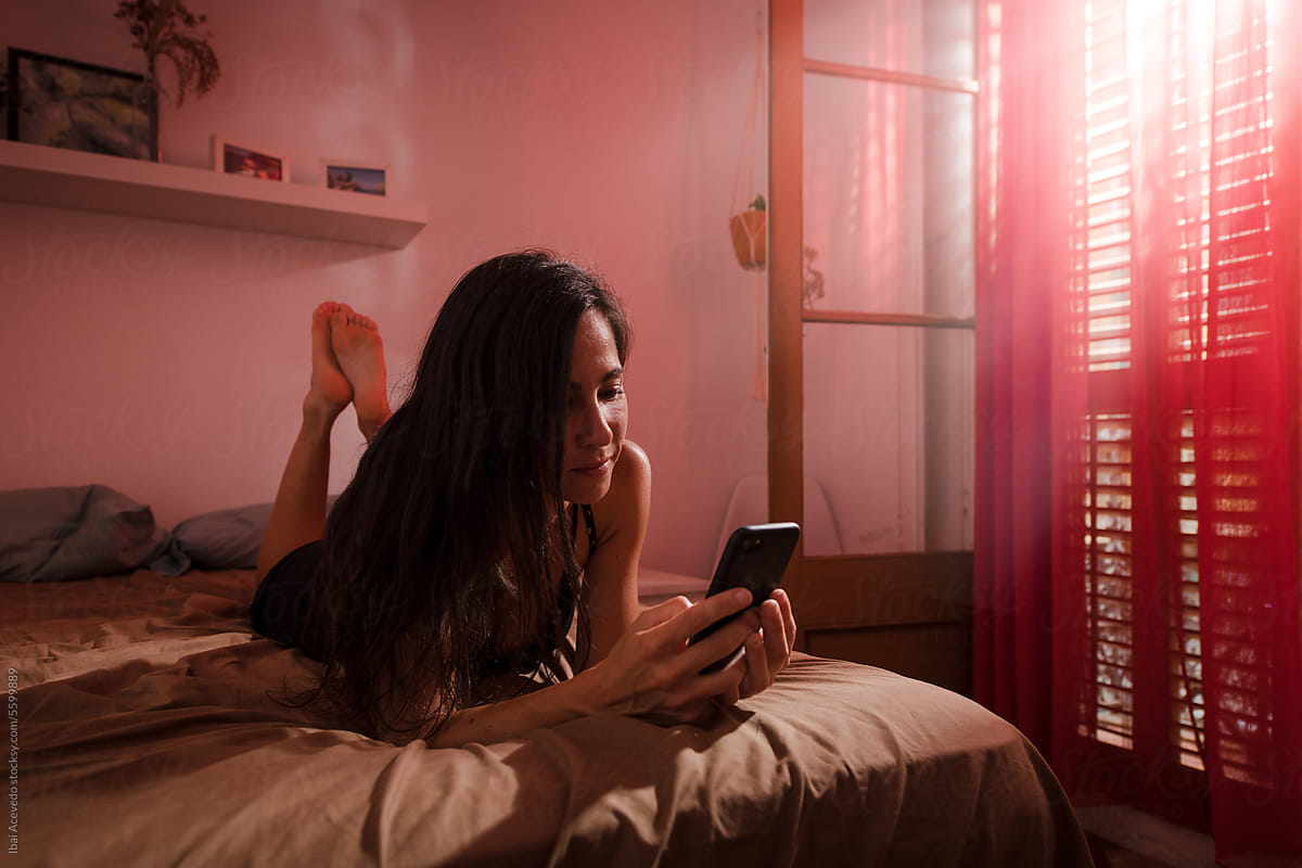 Amused woman using phone on bed during bright morning