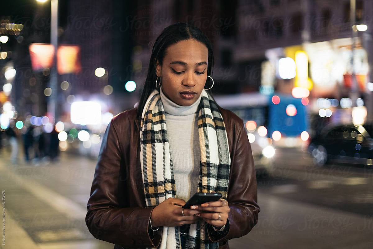 Stylish black woman texting with smartphone in the city at night