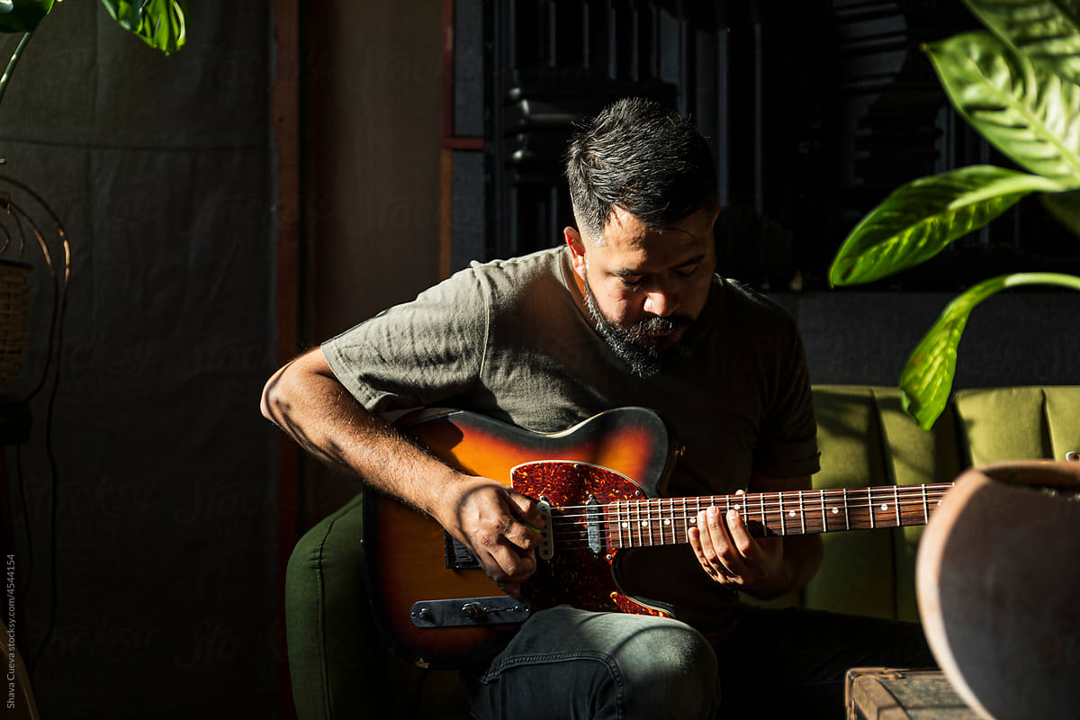 Man with beard playing an electric guitar in a  dark room with plants