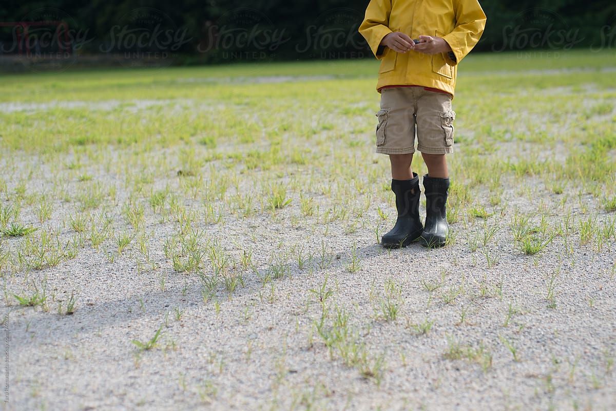 preschool boy stands in a field wearing a yellow rain jacket and boots