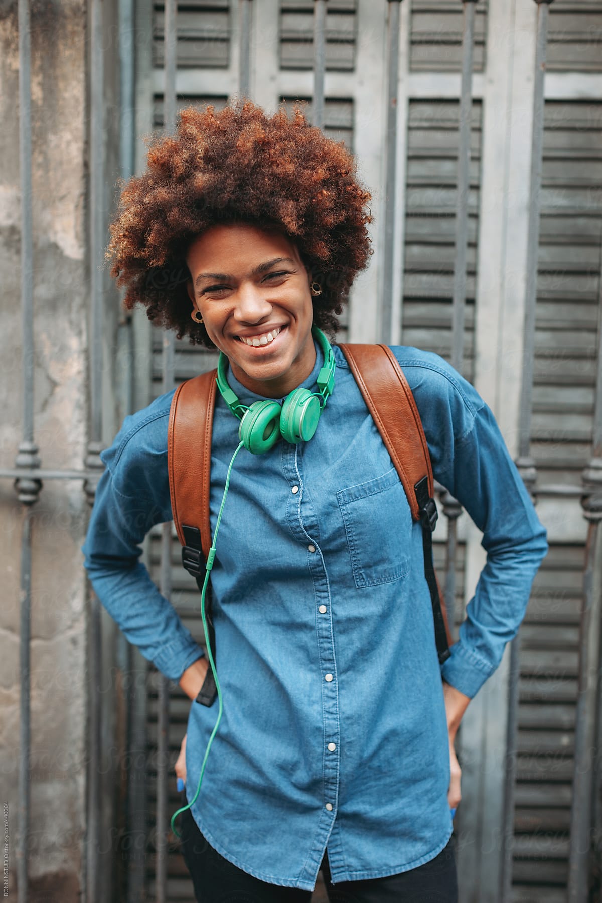 Afro american woman with leather backpack and green headphones smiling in outdoors.
