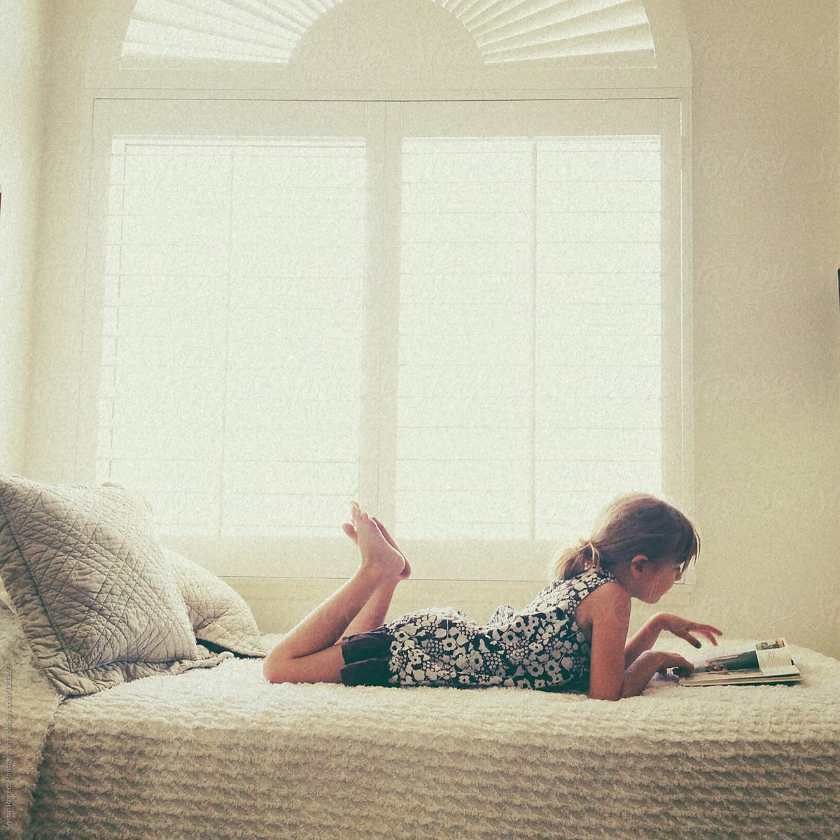 young girl reading book on bed next to window