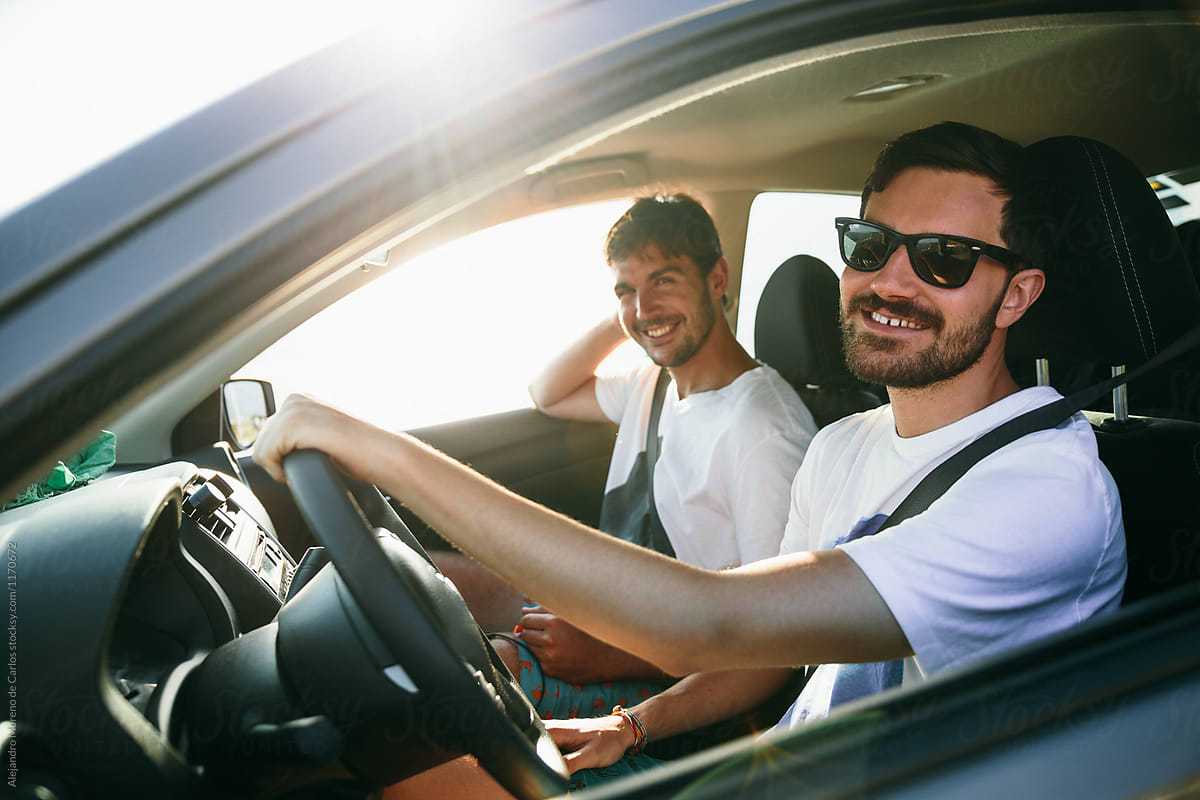 Two young man friends driving a car at sunset looking at camera during a road trip