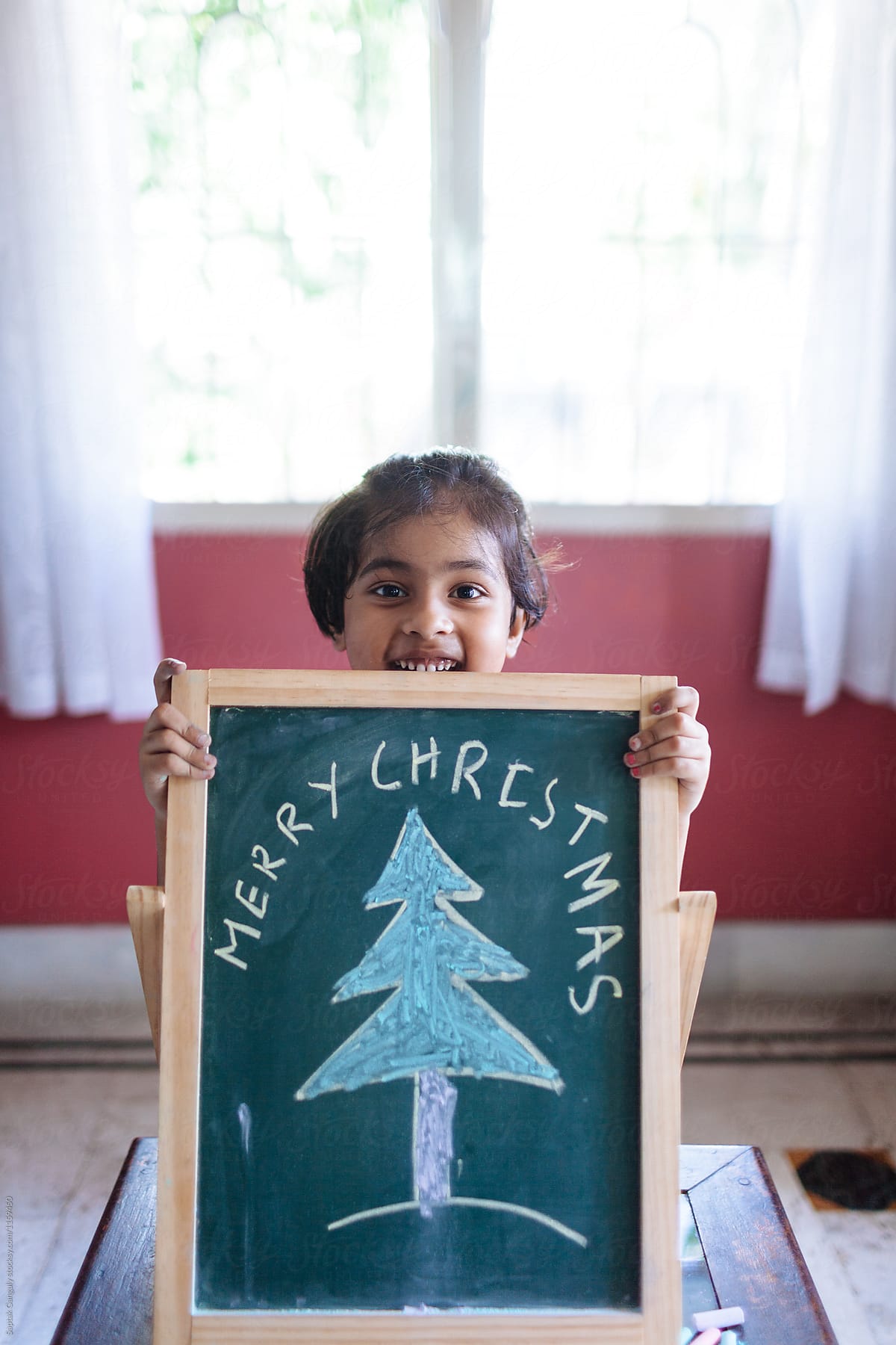 Little girl holding a green chalkboard with Merry Christmas and a Christmas tree drawn by her