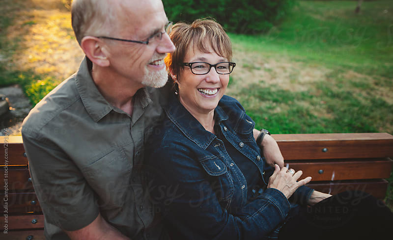 Happy Middle Aged Retired Couple Laughing And Cuddling Outside Sitting On A Bench By Rob And