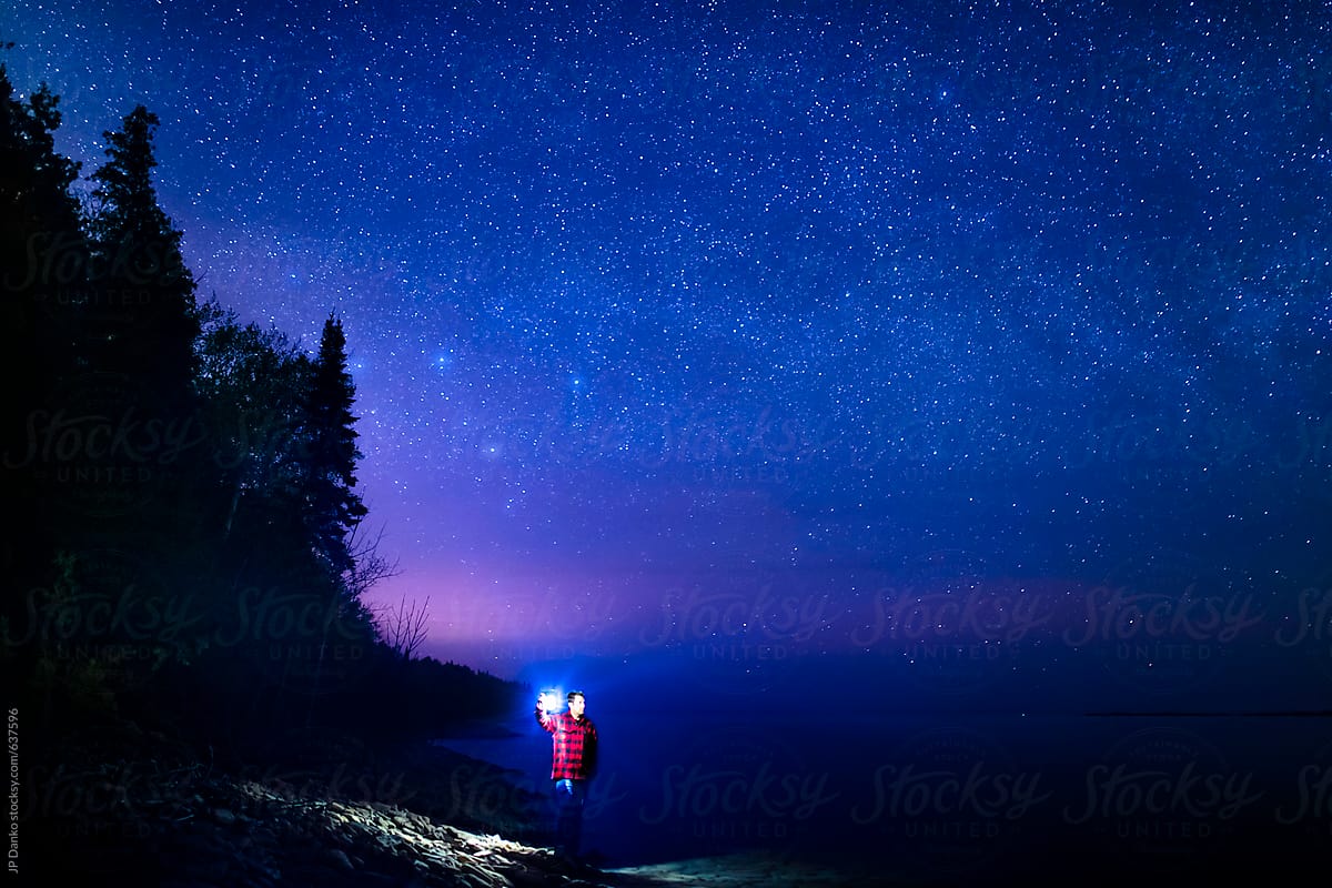 Man Illuminated By Flashlight Lantern Camping On Northern Beach Under Milkyway Stars