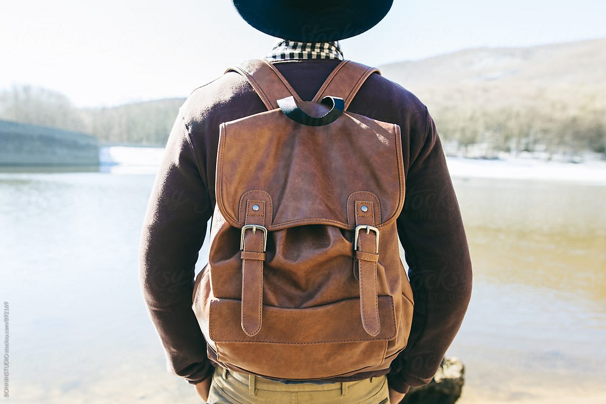 Back view of a mountaineer wearing a leather backpack in front of a lake.