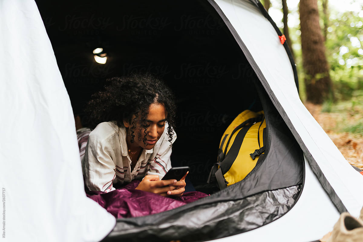 curly hair woman relaxing inside of a tent using her phone.