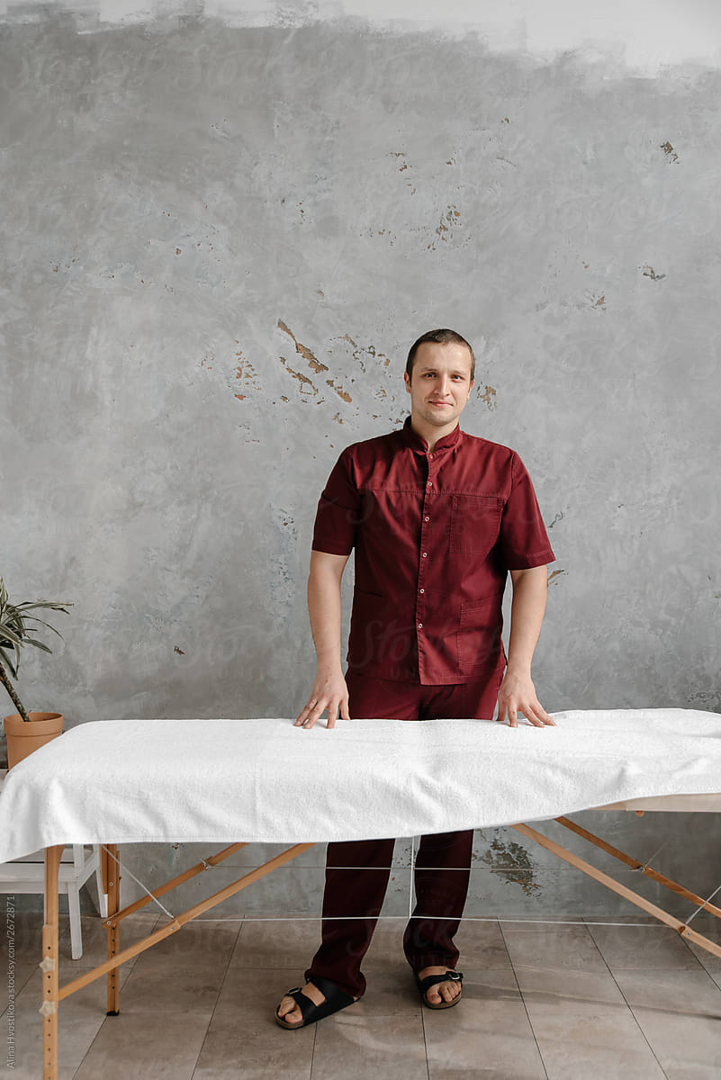 Male masseur wearing red uniform standing near table.