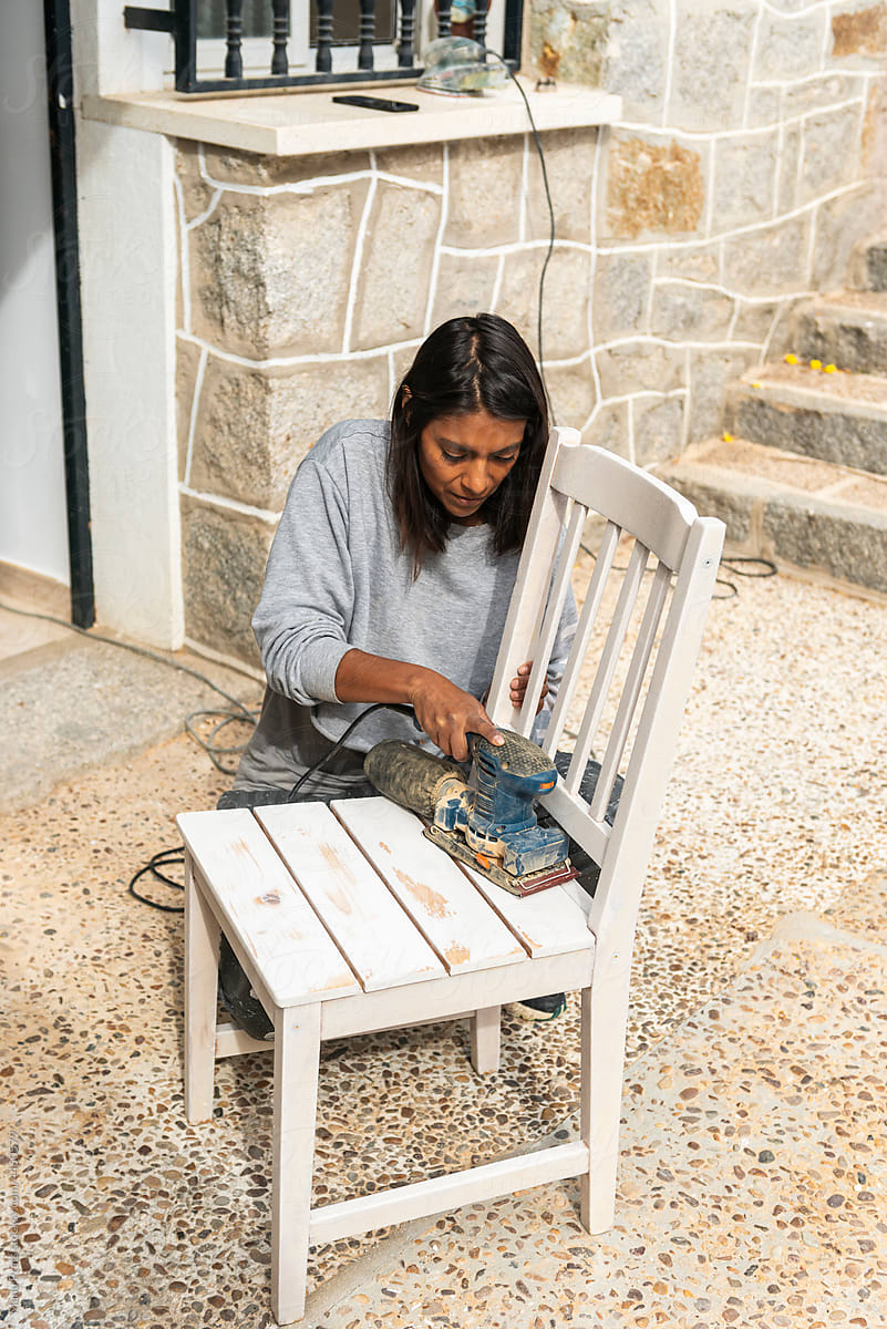 Brown skinned woman using sanding machine to restore a chair at home