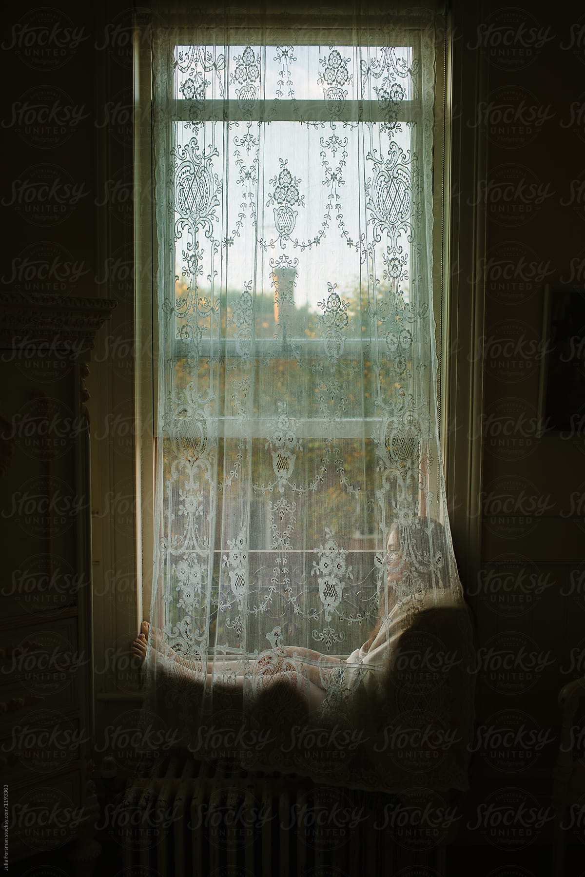 Woman sits behind a lace curtain at a window lost in her own thoughts.