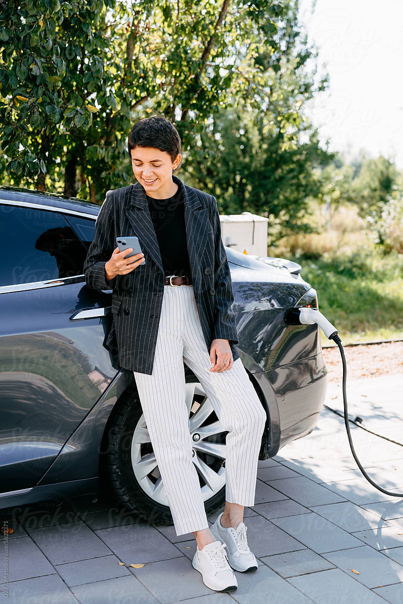 A woman uses a mobile phone standing near an electric car
