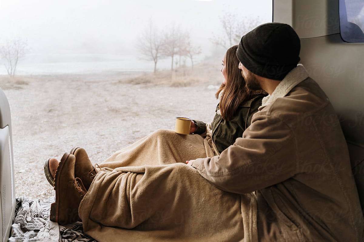 Young couple in a camper van in nature during winter having warm coffe looking away