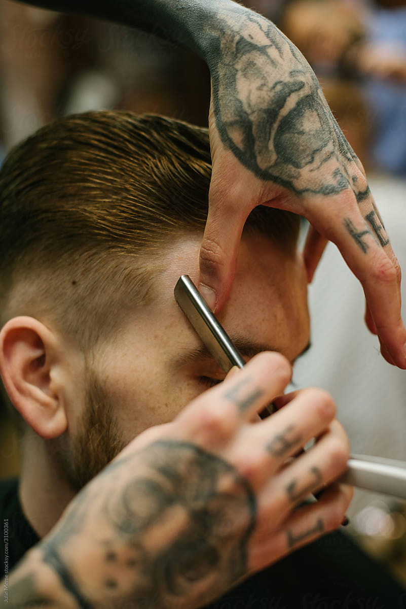 Closeup photo of a barber’s tattooed hands Shaving a ginger haired guy with a razor