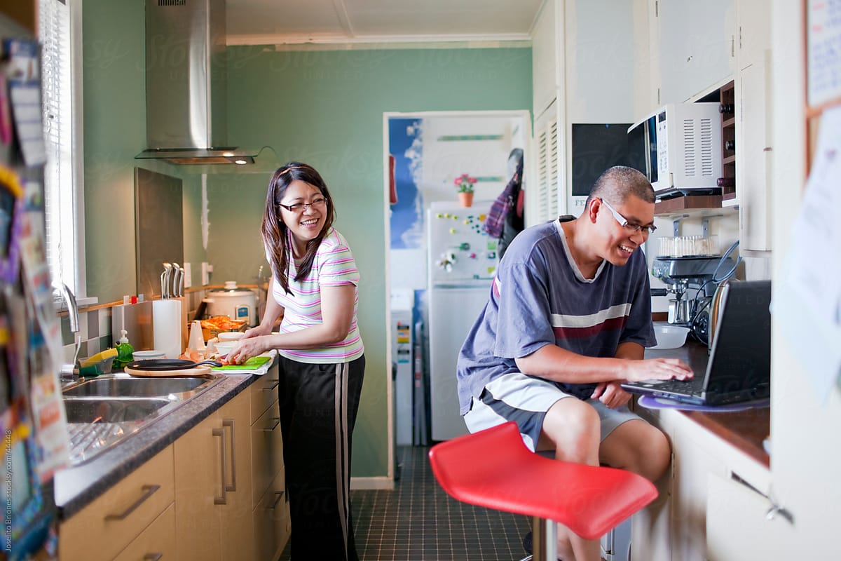 Husband Using Laptop And Wife Cooking In Kitchen Together Del 