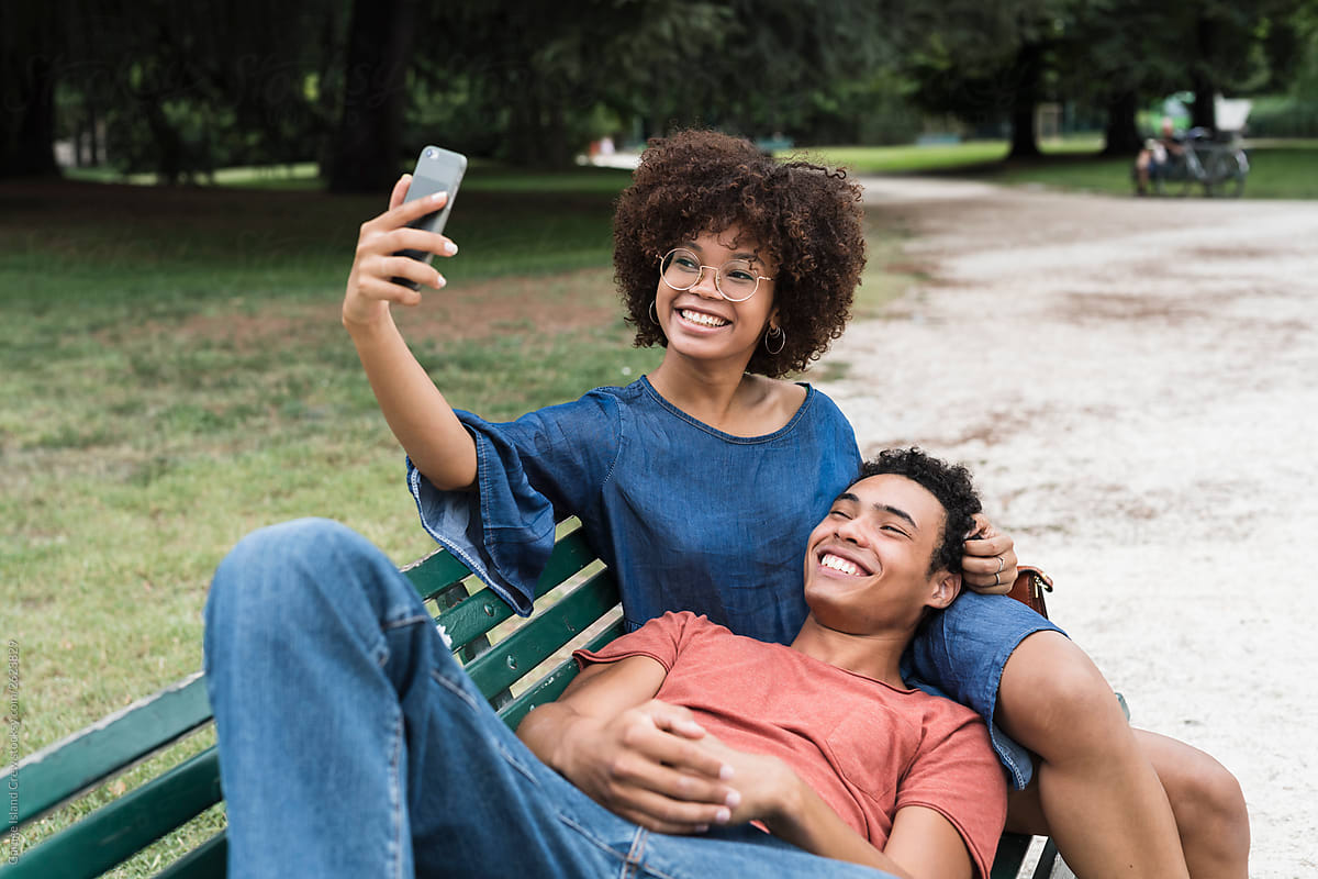 Happy young black couple sitting on the bench at the park taking