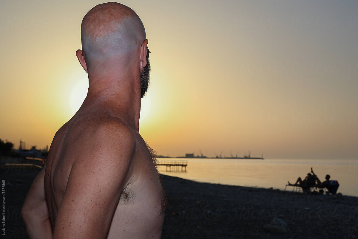 Naked bold man seen from a side looking the sun setting down in sea
