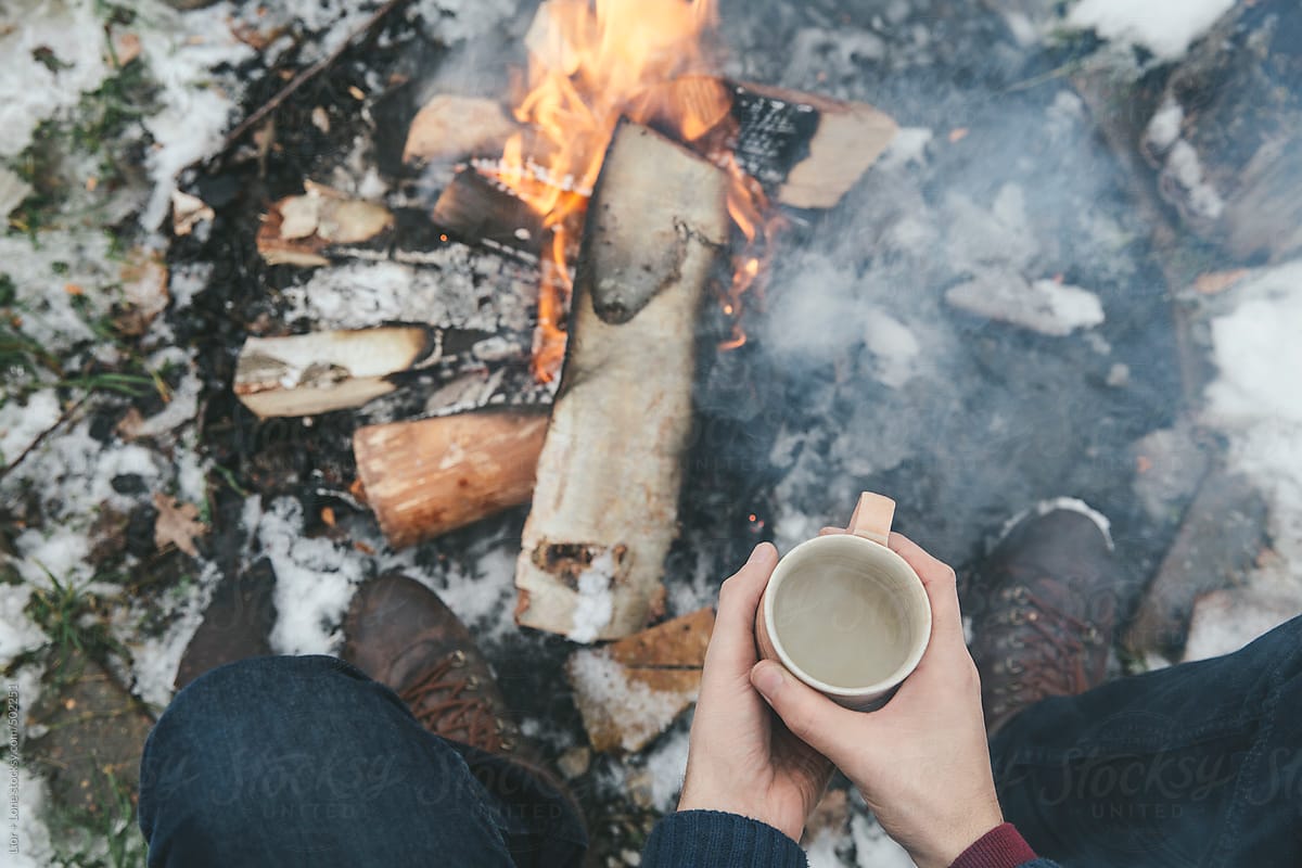 Hands holding a cup of warm tea next to bonfire in the snow