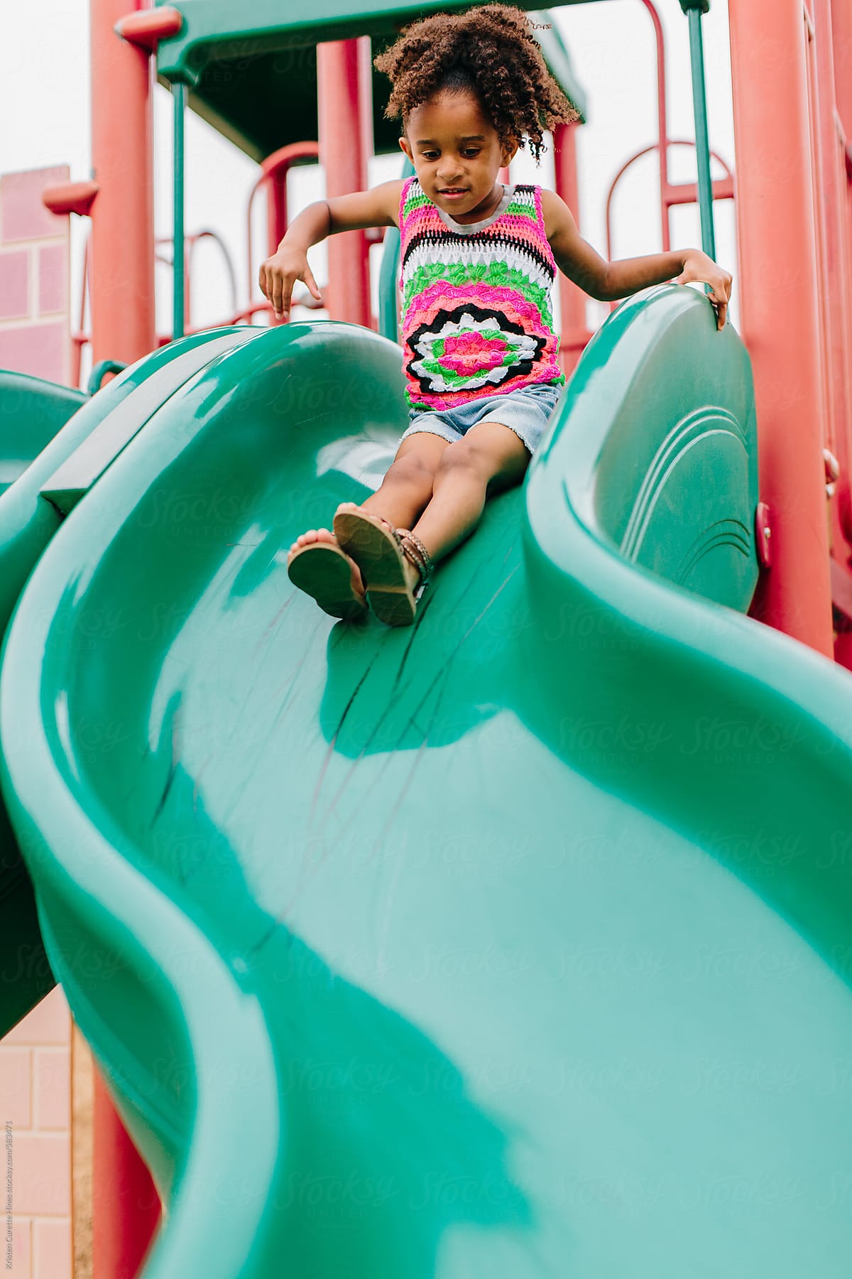 A Little Girl Sliding Down A Green Slide At The Playground. by