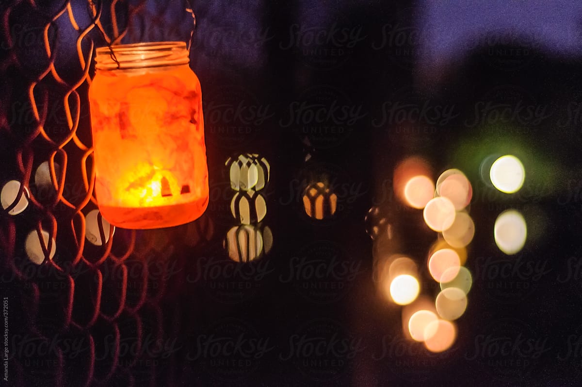 Mason jar with a lit candle  hanging on a fence at night.