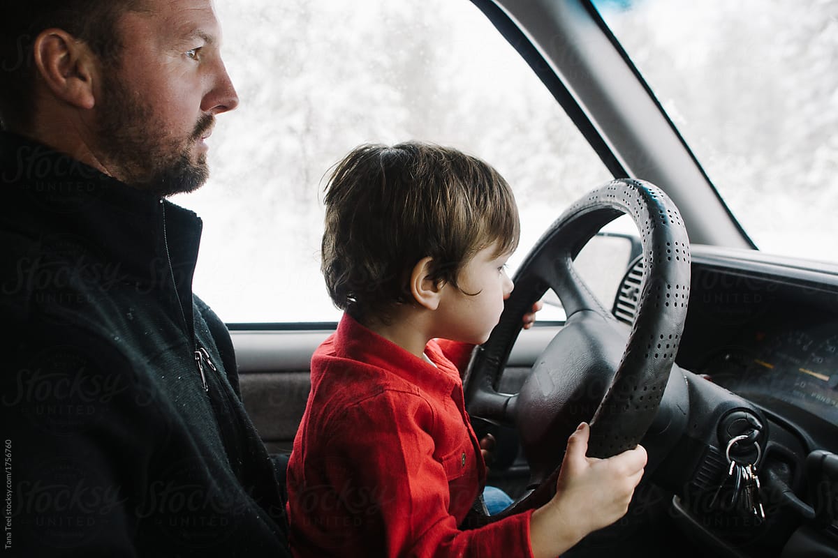 son sits on dad\'s lap helping drive while in open mountain area
