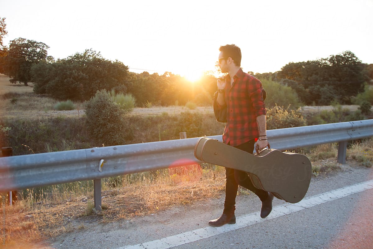 Man walking with his guitar and bag on the roadside at sunset